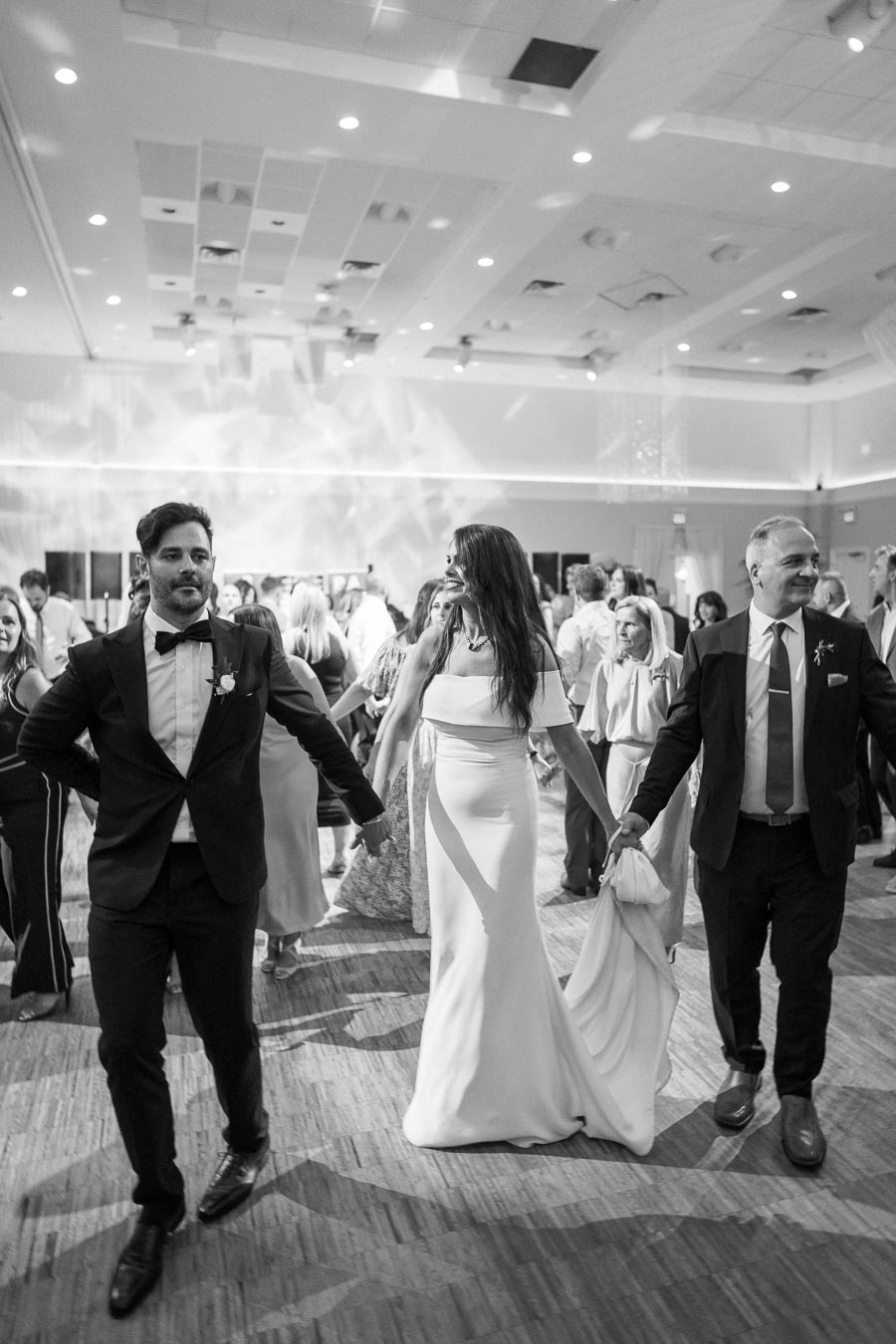 Black and white photo of a wedding reception with a bride in a white dress joyfully dancing with guests in a spacious, elegantly decorated hall.
