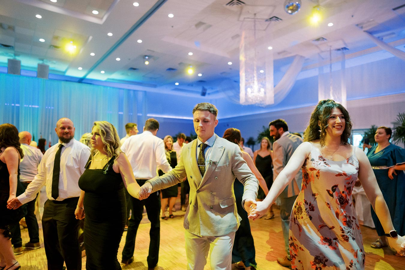 A group of people dressed in formal attire dance joyfully in a warmly lit ballroom, holding hands in a circular formation, creating a festive and celebratory atmosphere.
