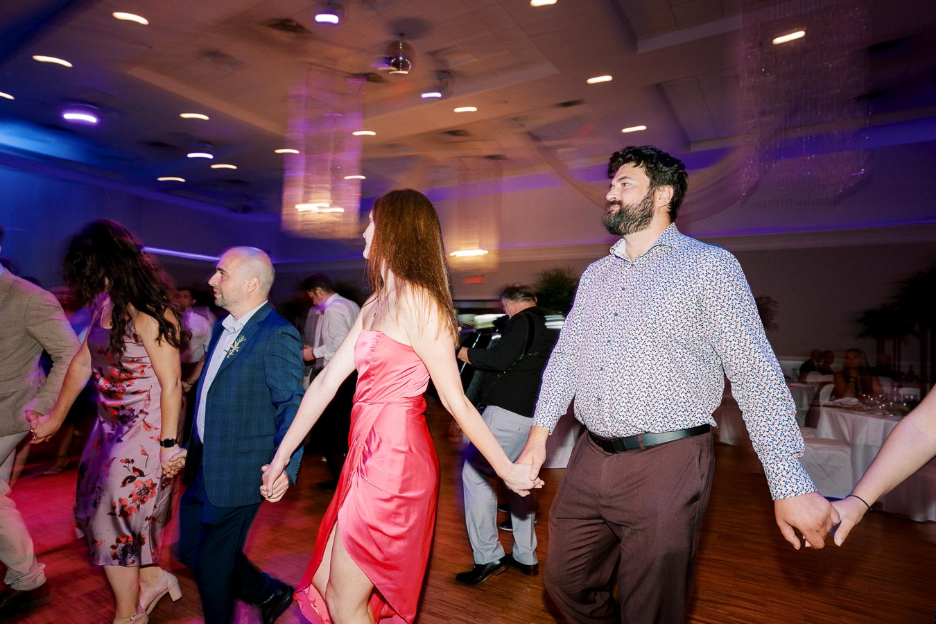 A group of people holding hands and dancing at a lively indoor event with colorful lighting and elegant decor.