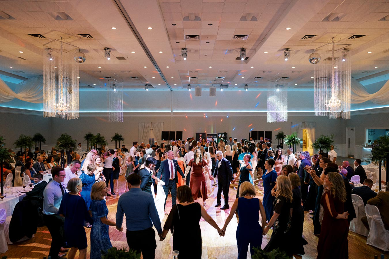 A lively wedding reception with guests dancing in a beautifully decorated ballroom, featuring elegant chandeliers, ambient lighting, and formal attire.