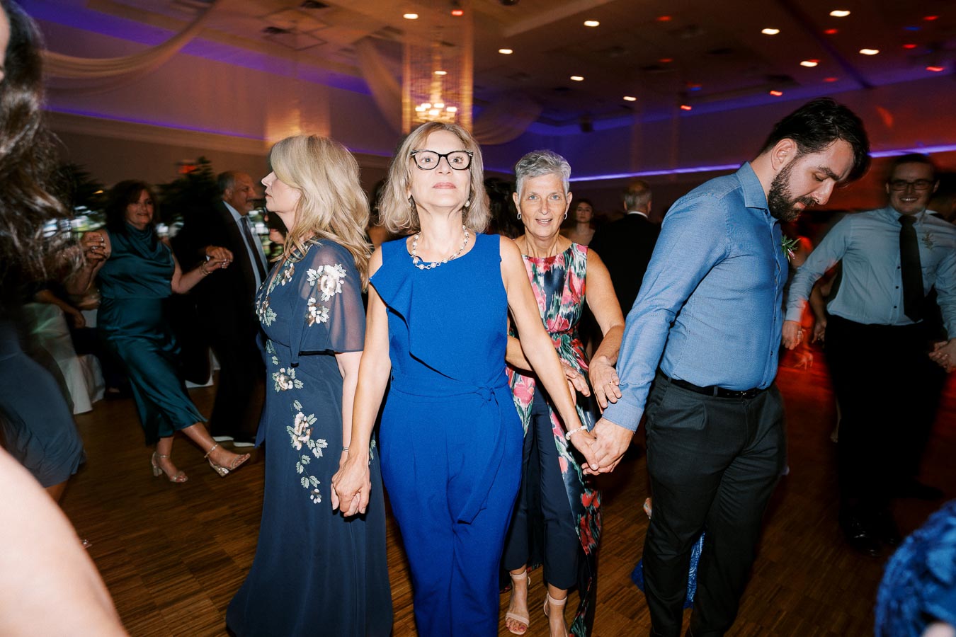 A group of people holding hands and dancing at a formal event, wearing elegant dresses and shirts, in a warmly lit banquet hall with draped ceilings and colorful lights.