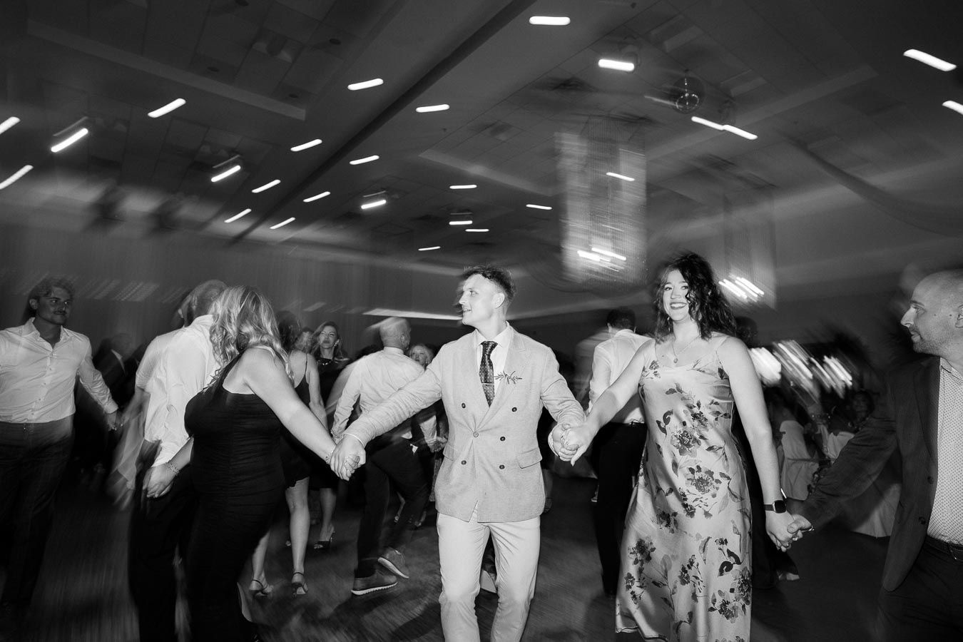A group of people joyously dancing in a circle at a lively celebration, captured in dynamic black and white motion.