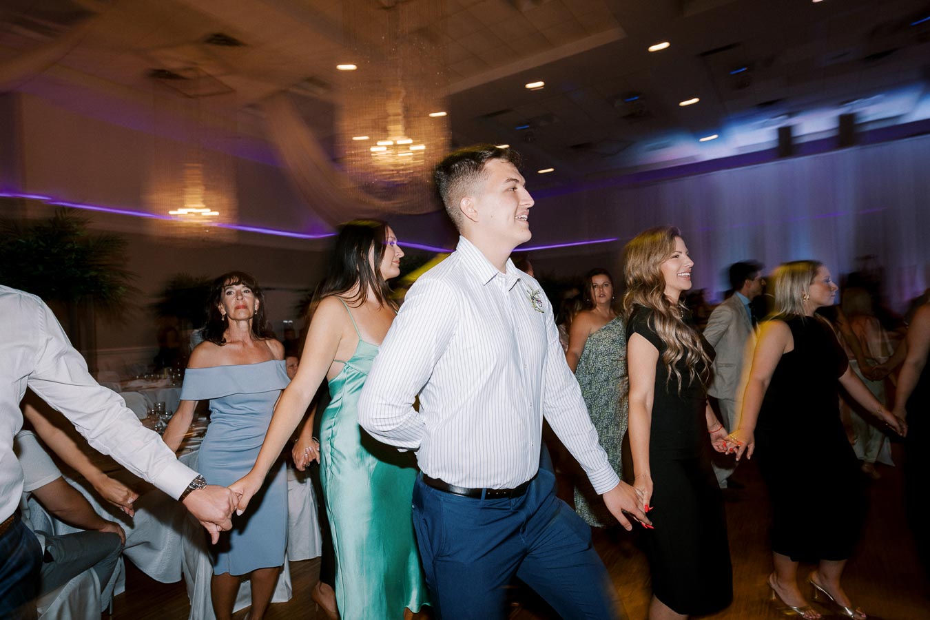 A group of people joyfully dancing at a wedding reception in an elegant ballroom setting, with chandeliers and ambient lighting enhancing the festive atmosphere.