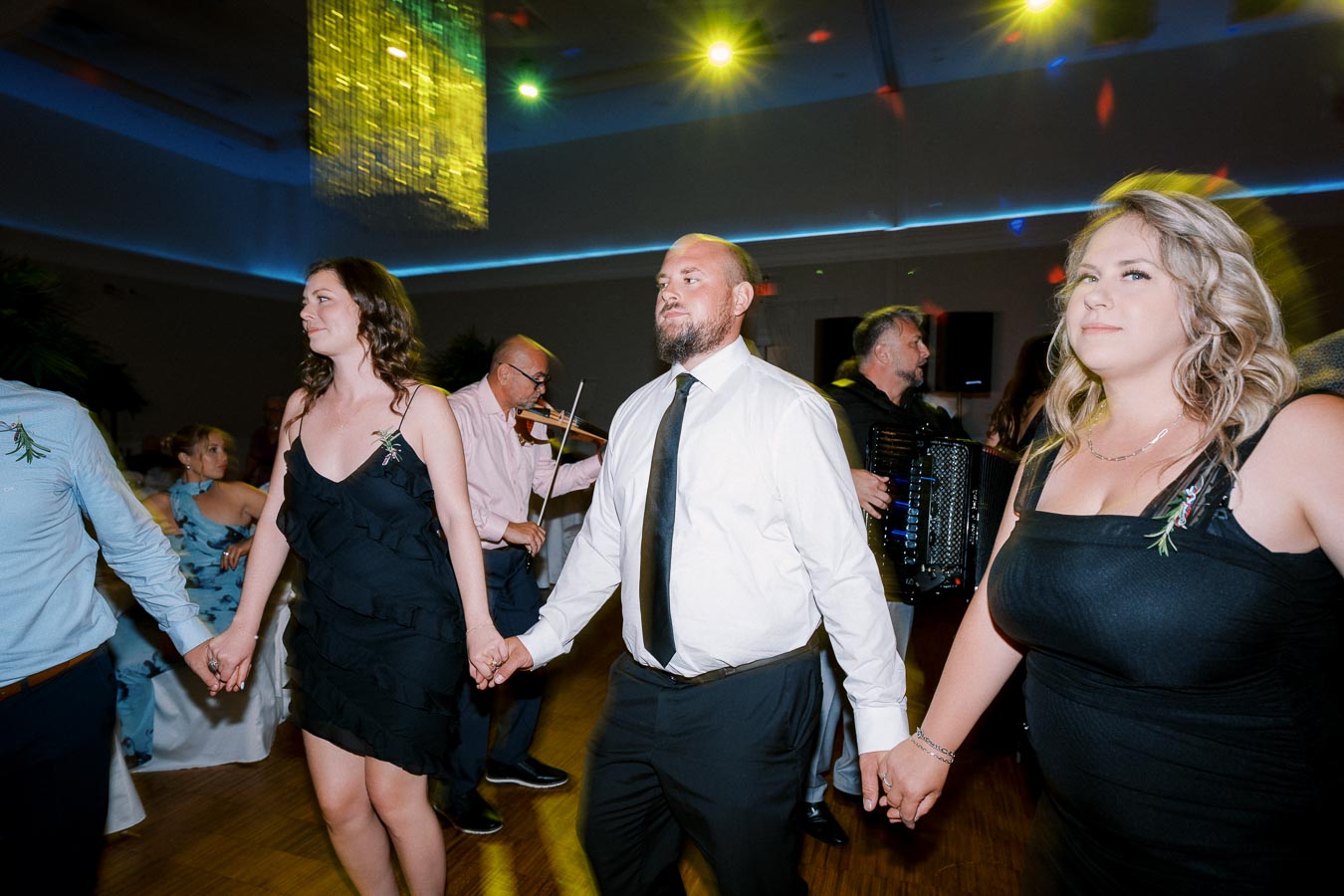 A group of people holding hands and dancing at an indoor celebration, with musicians playing violin and accordion in the background. The room is decorated with colorful lights creating a festive atmosphere.