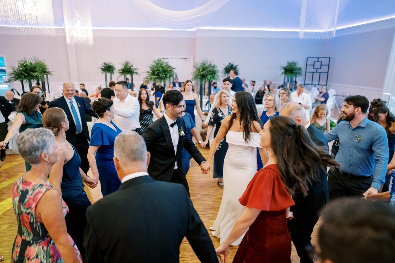 Guests enjoying a lively dance at a wedding reception, with elegantly dressed attendees holding hands in a circle on a polished wooden floor, surrounded by greenery and soft lighting.
