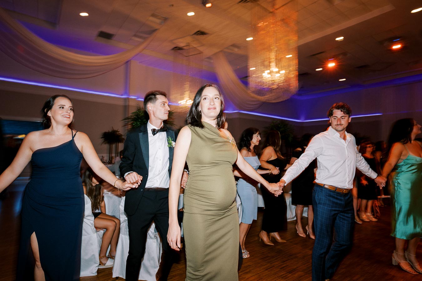 A group of elegantly dressed people holding hands while dancing at a formal event, indoor setting with dim lighting and a chandelier overhead.