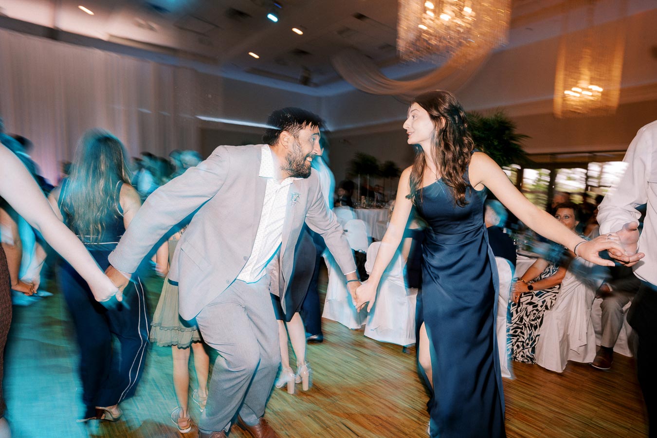A joyful wedding reception dance floor scene with a man in a gray suit and a woman in a navy blue dress dancing energetically, surrounded by other guests in a warmly lit ballroom setting.