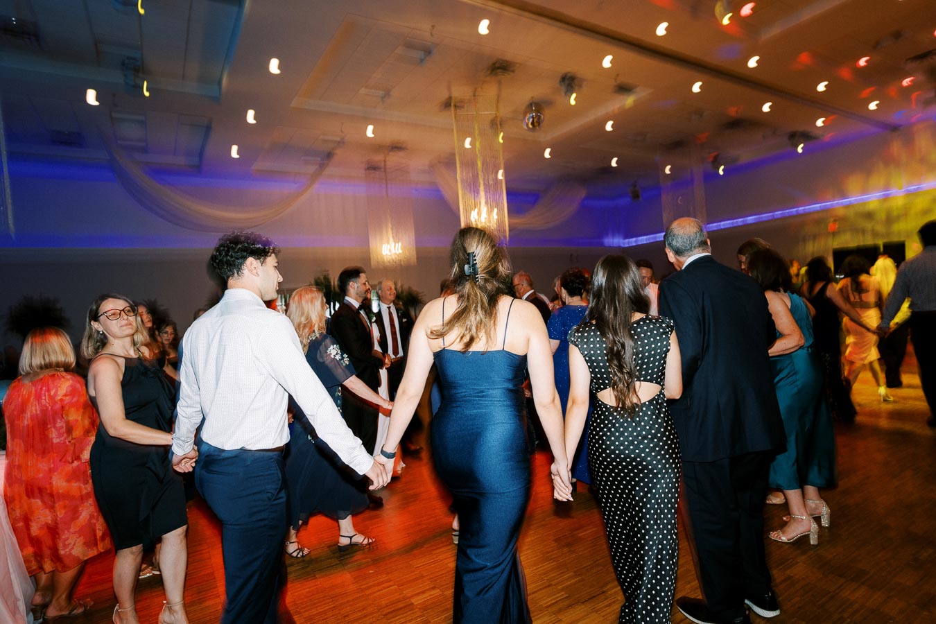 A lively ballroom dance event with elegantly dressed guests holding hands on a wooden dance floor, under colorful lighting in a decorated venue.