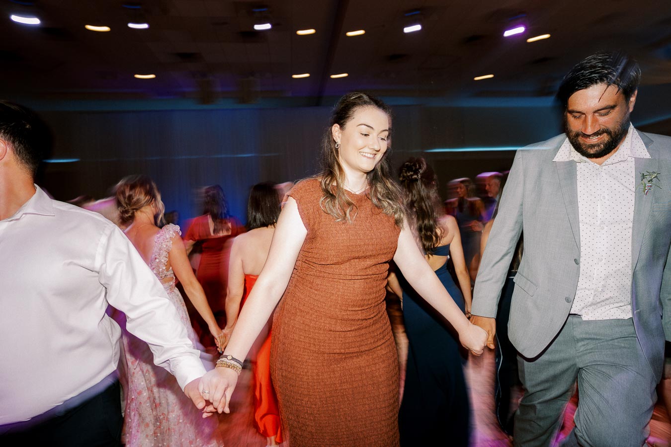 People dancing joyfully at a lively party, wearing formal attire, under colorful lights in a reception hall.