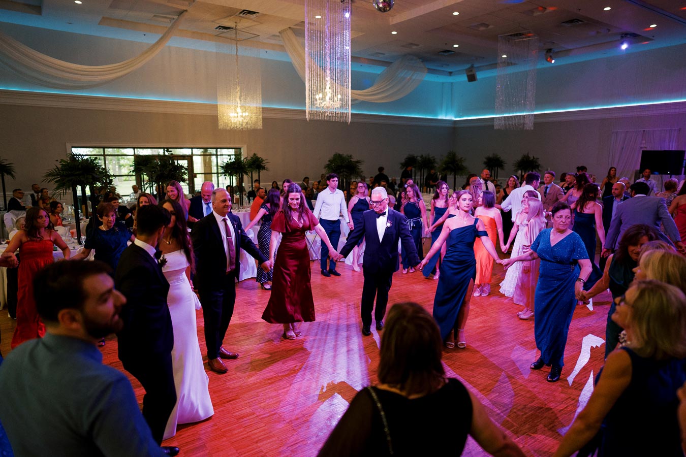 A lively wedding reception with guests in formal attire dancing in a circle under elegant chandeliers and draped ceilings in a beautifully decorated hall.
