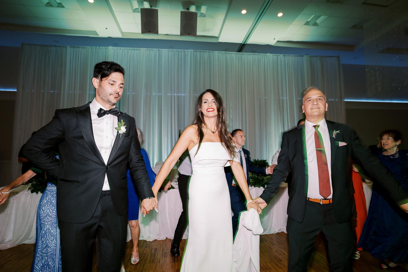 A bride in a white dress holding hands with two men in suits, dancing joyfully at a wedding reception. The venue features elegant lighting and draped curtains, enhancing the celebratory atmosphere.