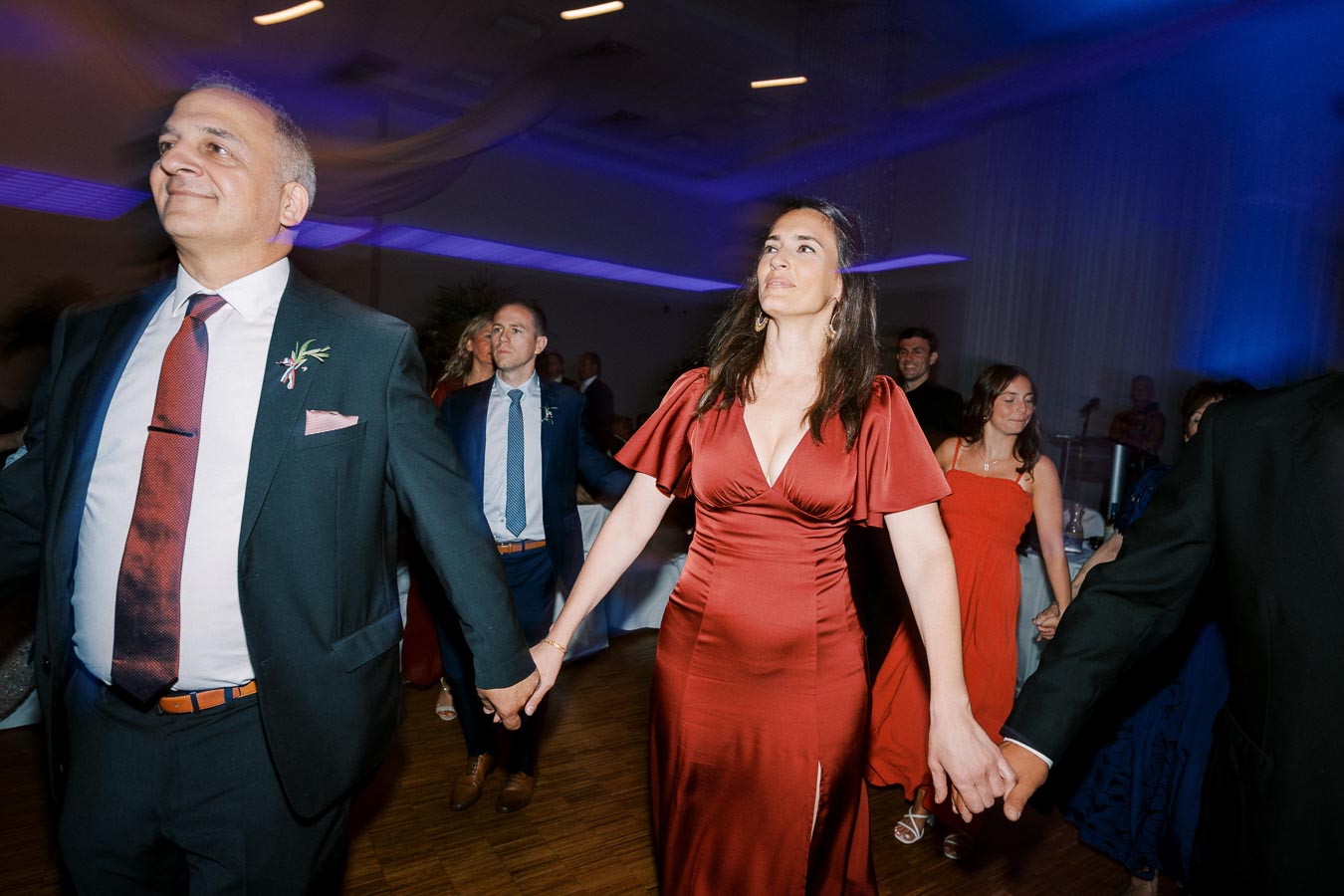 Guests dancing and holding hands at a lively wedding reception, with ambient blue lighting in the background, capturing the joyful atmosphere and stylish attire.