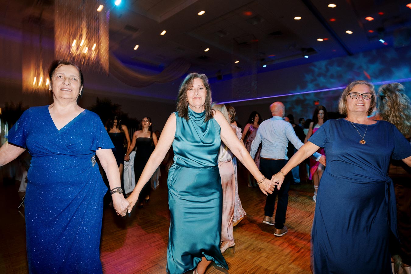 Three women in elegant dresses are joyfully dancing and holding hands at a lively indoor event, with colorful lights and other partygoers in the background.