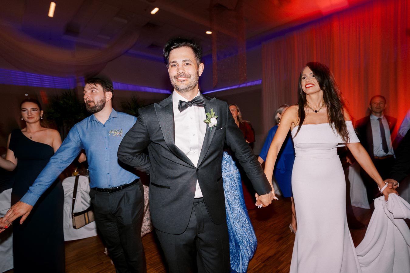 A joyful wedding celebration with guests on a dance floor. Centered is a man in a tuxedo and boutonniere, joined by a smiling woman in a white gown. The background features a warmly lit venue with other attendees enjoying the festivities.