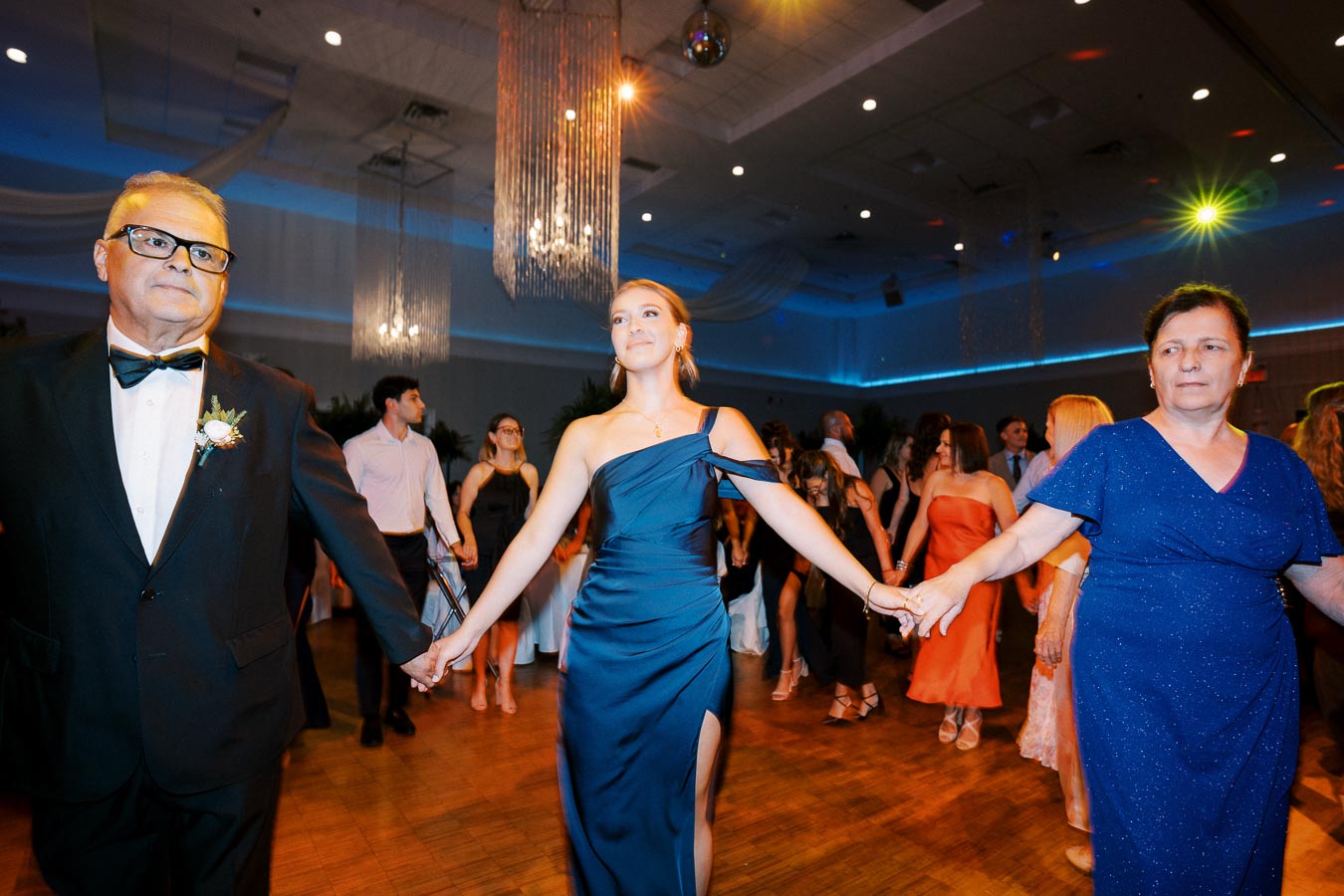 A group of people holding hands and dancing at an elegant evening event, with women in formal dresses and a man in a tuxedo, creating a festive and joyous atmosphere in a beautifully decorated venue with chandeliers.