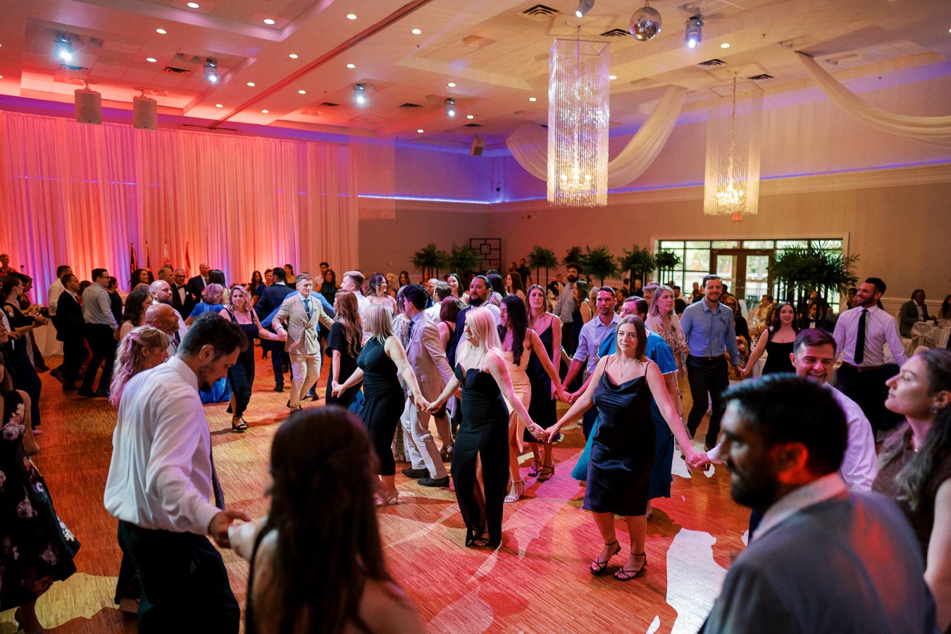 A lively wedding reception with guests joyfully dancing in a circle on a beautifully lit wooden dance floor, surrounded by elegant drapery and hanging chandeliers.