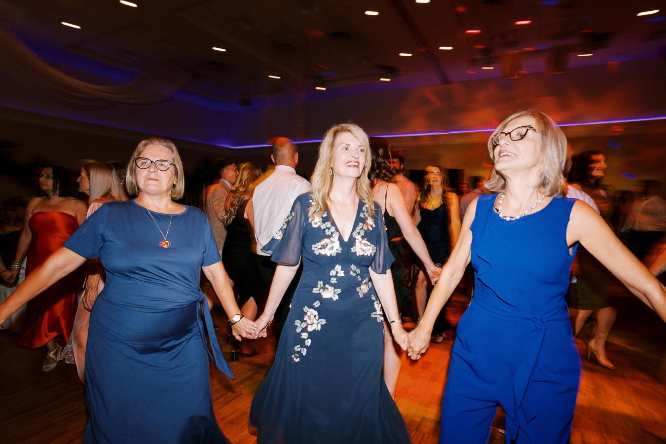 Women joyfully dancing and holding hands at a festive indoor event with colorful lighting and a lively atmosphere.