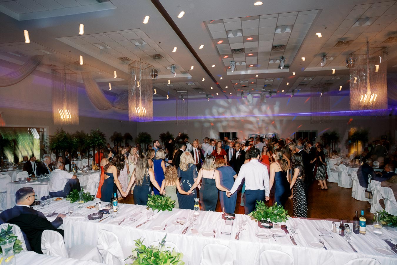 A large group of people dancing in a decorated banquet hall with elegant lighting and well-set tables, capturing the vibrant atmosphere of a formal event or celebration.