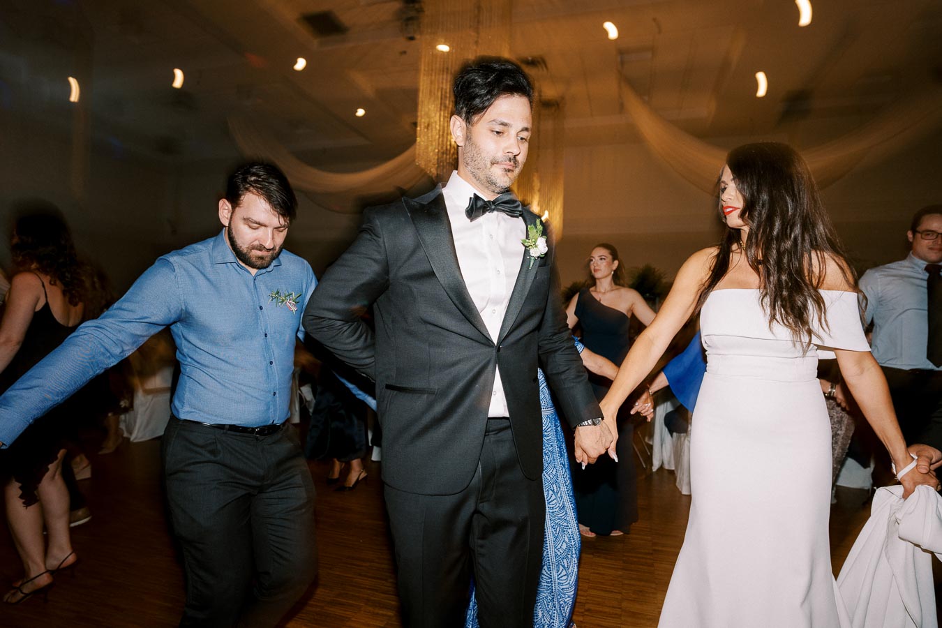 A group of elegantly dressed people holding hands and dancing at a formal event, with a man in a black tuxedo and a woman in a white dress leading the dance. The setting appears to be a reception hall with decorative lighting.