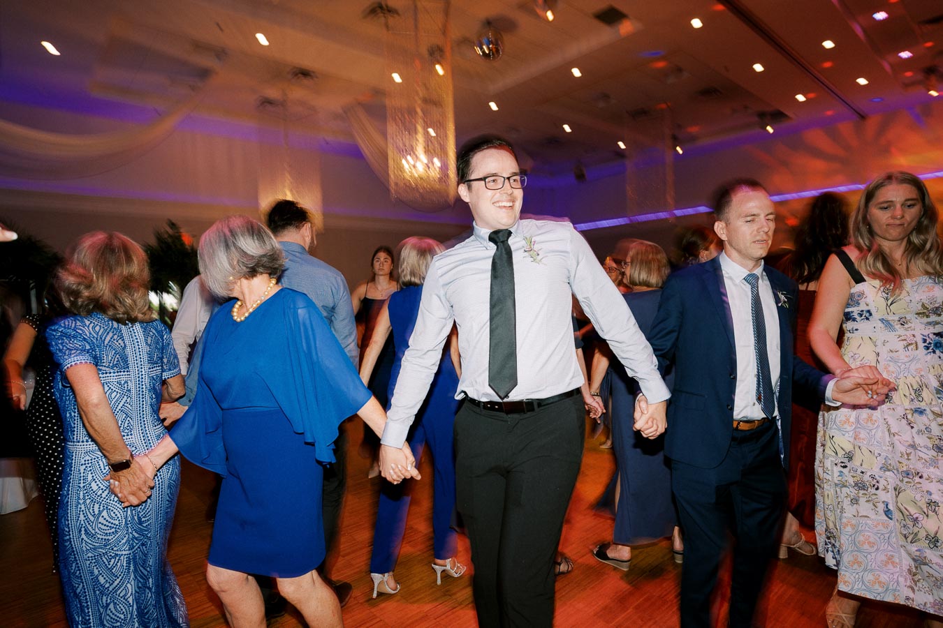 A group of people joyfully dancing at a lively indoor event, with colorful lighting and elegant attire, creating a vibrant atmosphere.