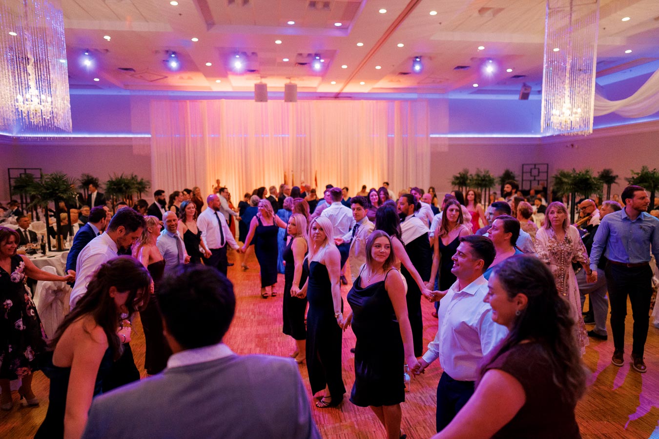A lively wedding reception with many guests dancing in a beautifully decorated ballroom, featuring soft lighting and elegant chandeliers.