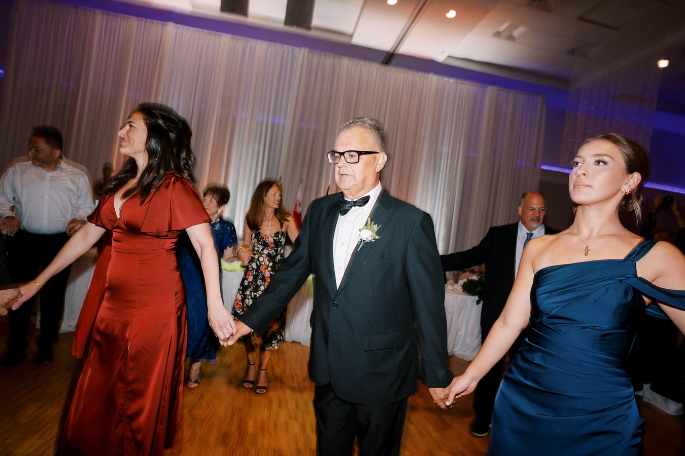 People holding hands and dancing in a formal event, featuring individuals in elegant dresses and suits, with a decorated venue background.