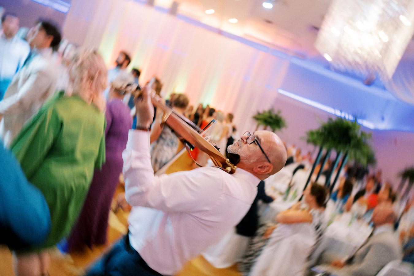 A musician energetically plays the violin in a lively wedding reception, surrounded by dancing guests in colorful attire, under elegant chandeliers and ambient lighting.