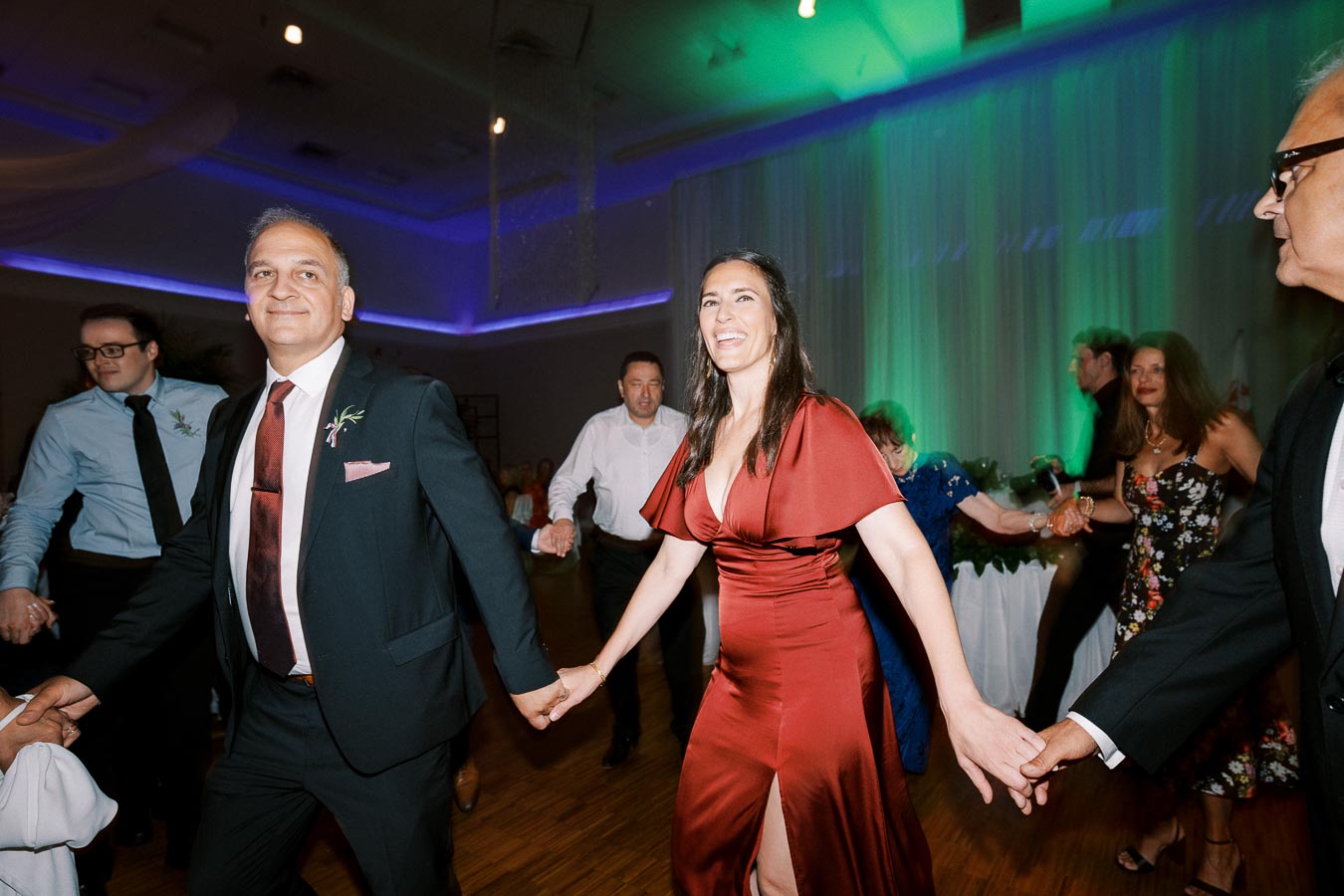 A group of people joyfully dancing in a well-lit ballroom with colorful lighting, featuring a woman in a red dress and a man in a suit holding hands, capturing a lively and festive celebration atmosphere.