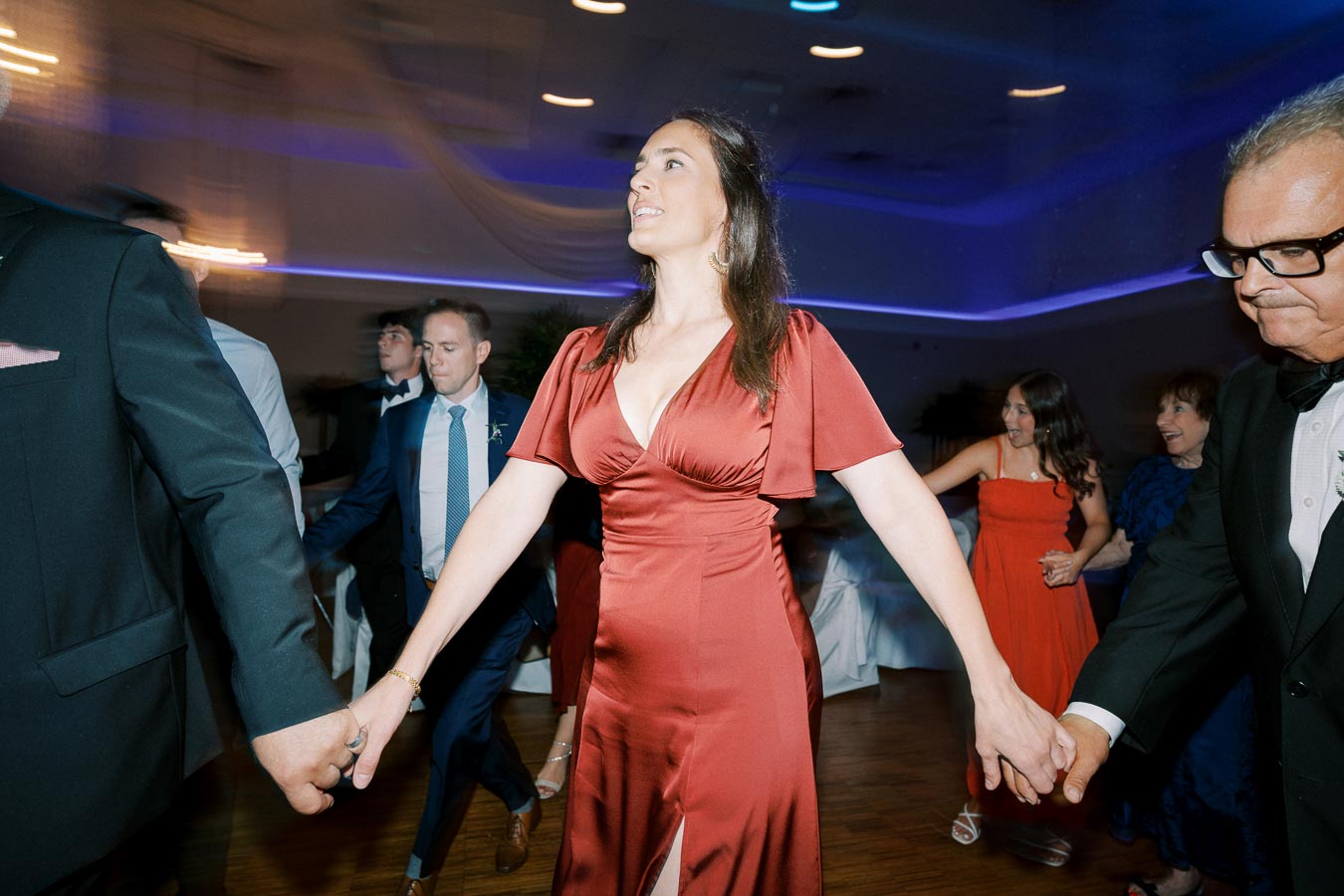 A group of people dancing in a warmly lit event space, with a woman in a red dress leading the circle, creating a joyful and lively atmosphere at a celebration or wedding.
