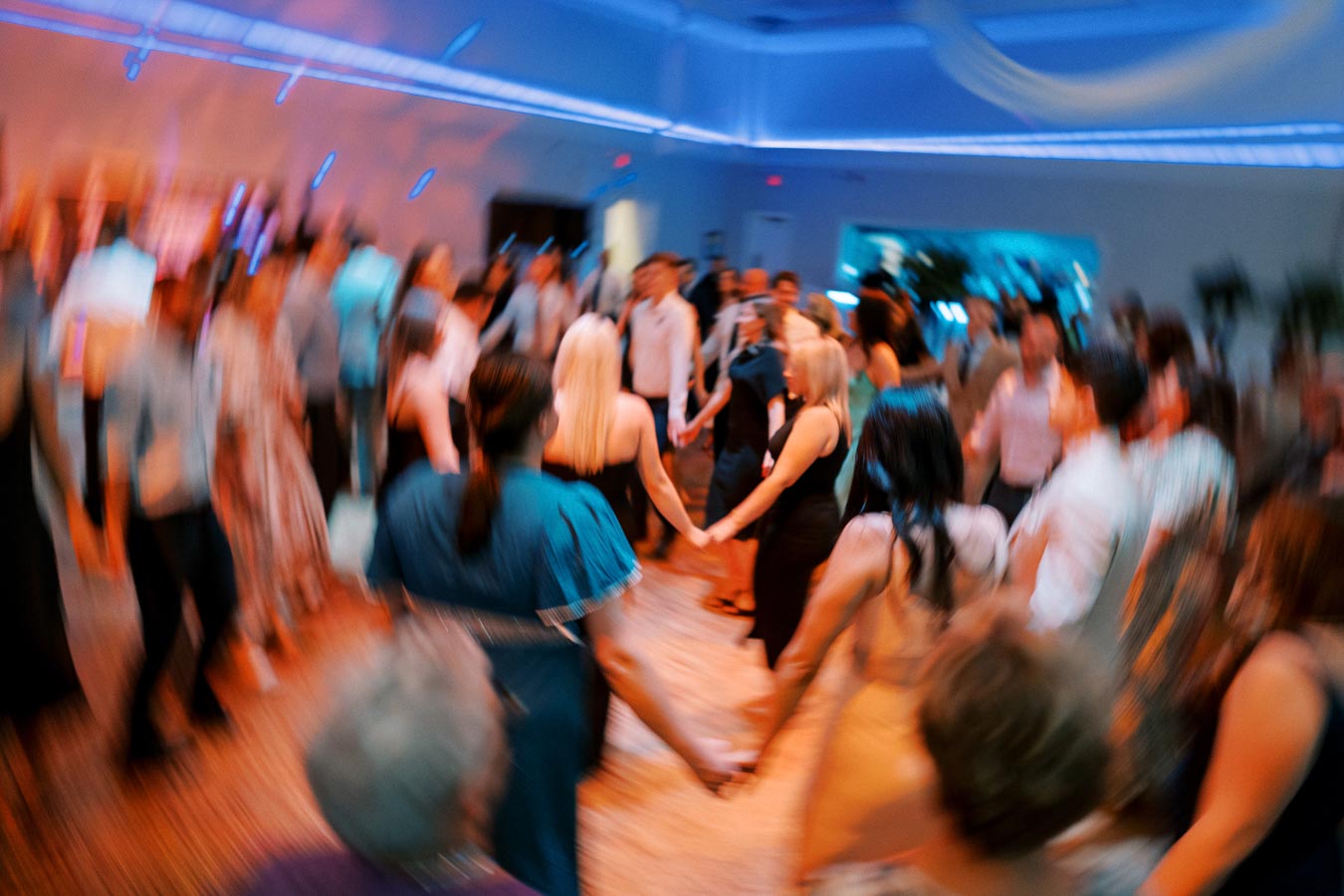 A group of people holding hands and dancing in a circle at a lively indoor event with colorful lighting and a blurred motion effect.