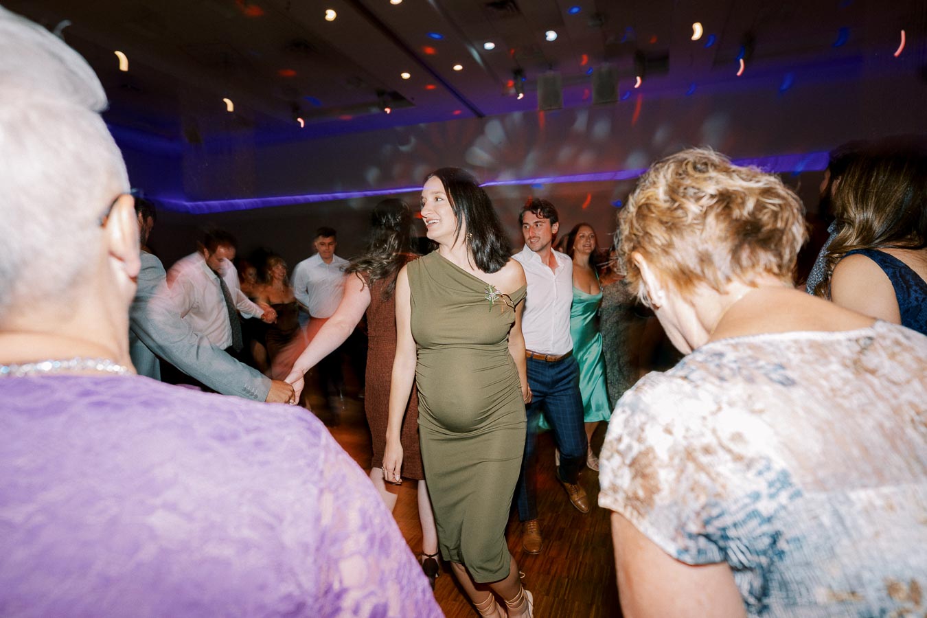 A lively dance floor scene featuring a group of people enjoying music at a social event, with a woman in a green dress smiling as she dances with others, under vibrant colored lights.