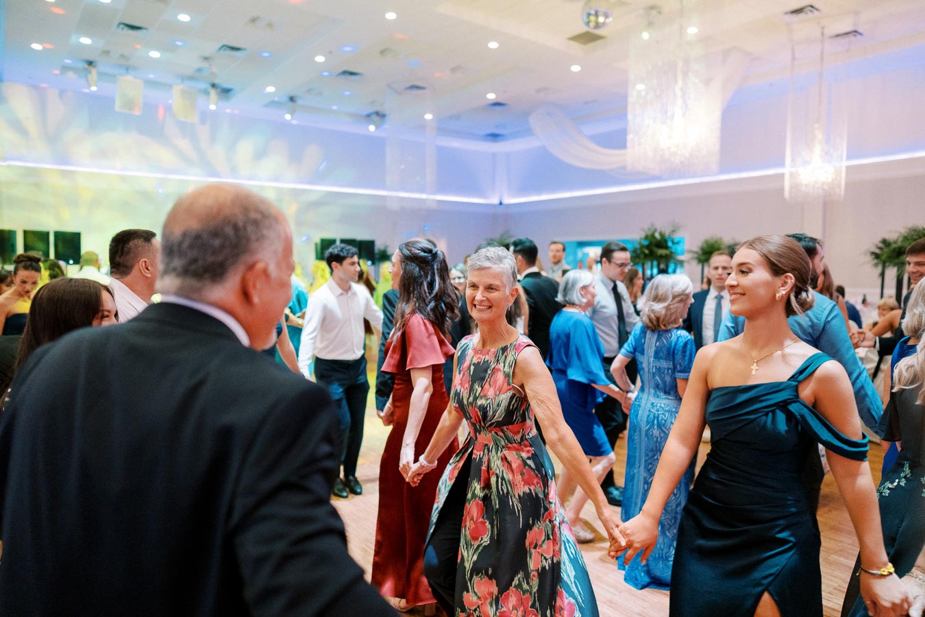 A joyful group of people dancing elegantly at a formal event in a beautifully decorated venue, highlighted by vibrant lighting and elegant attire.