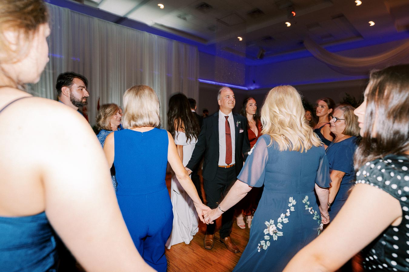 Group of people holding hands and dancing in a circle at an indoor event with soft lighting, wearing formal attire.