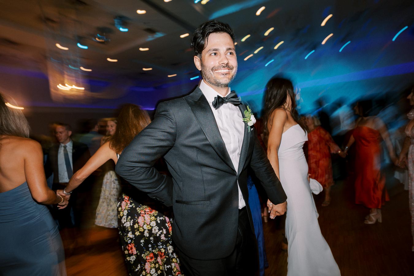 A man in a black tuxedo smiling while holding hands with a woman in a white dress at a lively wedding reception. The background is blurred with colorful lights and other guests dancing.