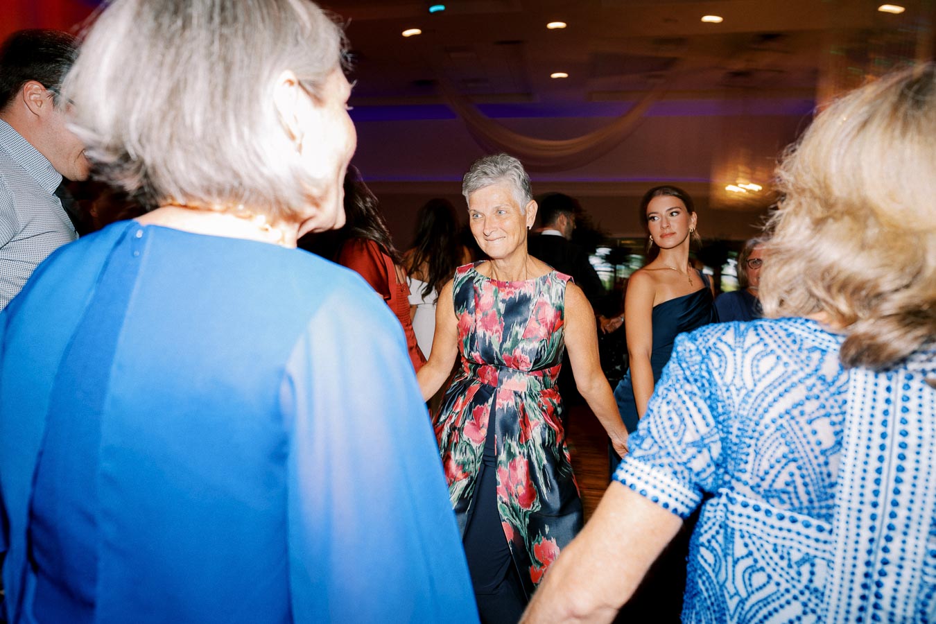 Senior woman dancing joyfully with friends at a vibrant party, wearing a floral dress, surrounded by guests in elegant attire.