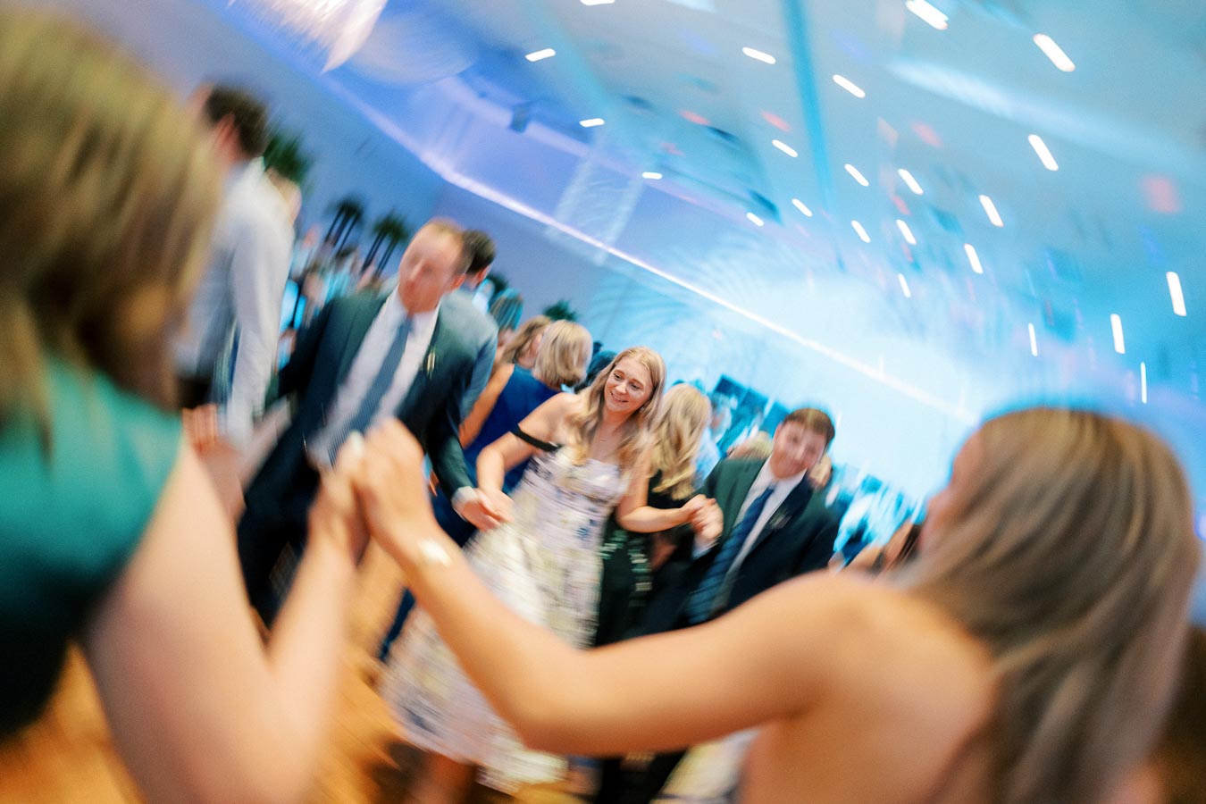 A lively group of people dancing at a wedding reception in a bright, elegantly decorated venue with blue lighting effects creating a festive atmosphere.