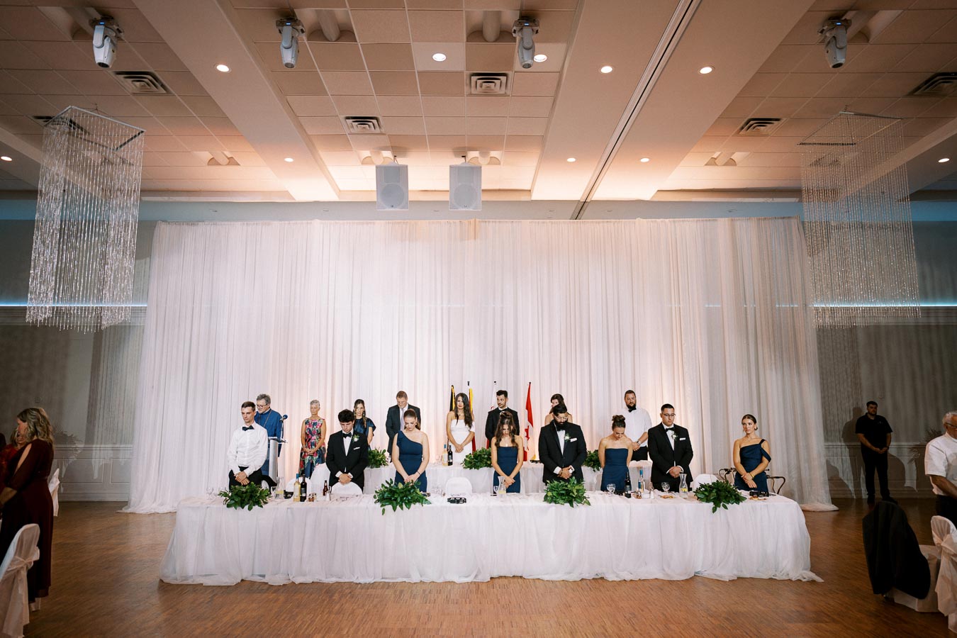 Elegant wedding reception with a long head table, adorned in white linens and greenery, where guests are standing in formal attire under a lavishly decorated ceiling, creating a sophisticated atmosphere.