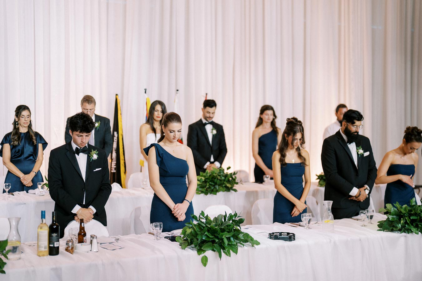 A group of elegantly dressed men and women, some in formal attire and others in blue dresses, stand solemnly behind a table adorned with greenery at a formal event, possibly during a moment of silence or prayer, with a backdrop of white drapery and flags.
