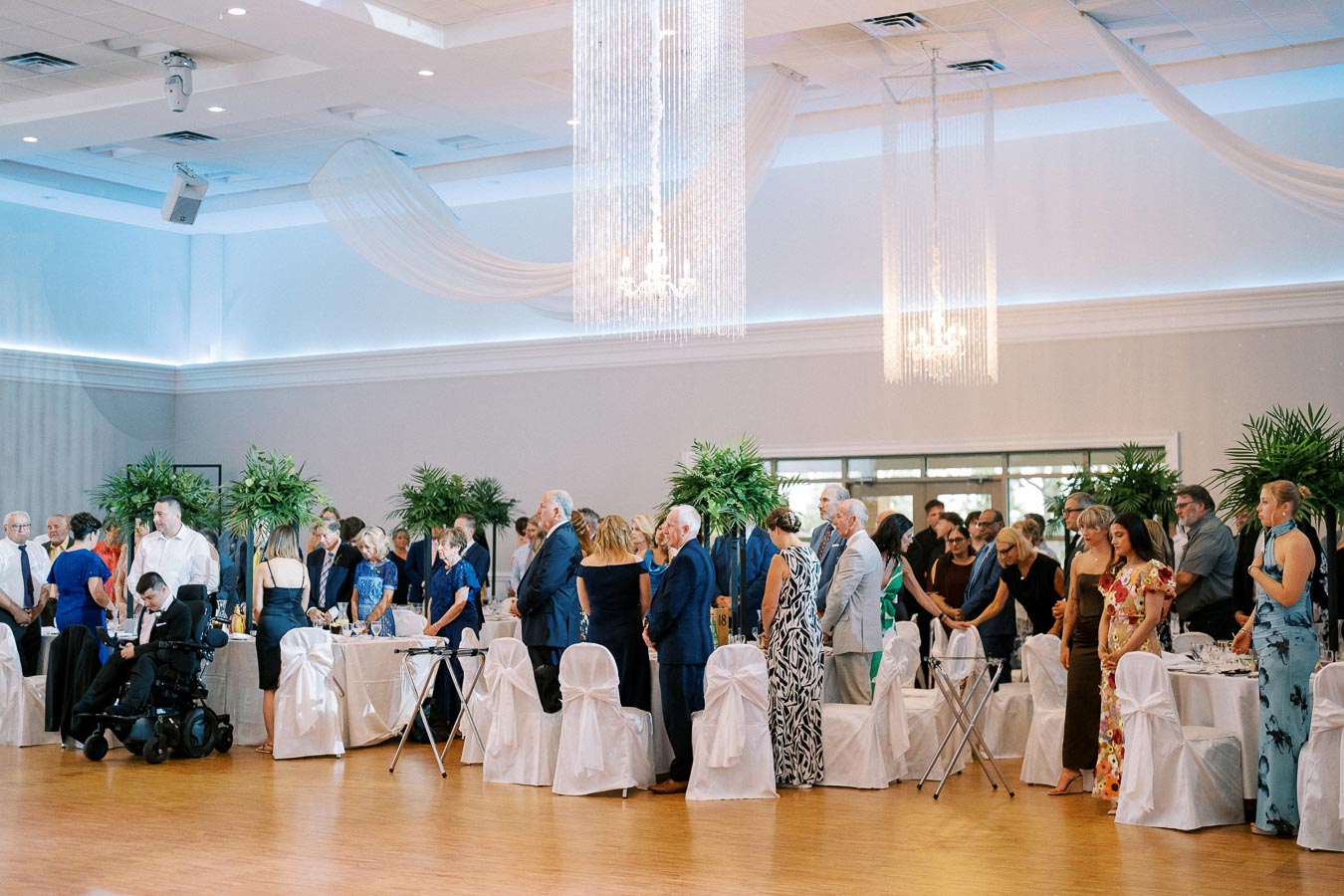 A large group of elegantly dressed guests standing in a beautifully decorated banquet hall with chandeliers and greenery, attending a formal event or celebration.