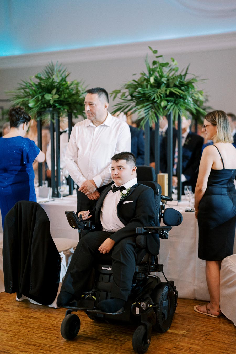 Formal event with a young man in a wheelchair wearing a suit and bow tie, surrounded by elegantly dressed guests and green floral arrangements.