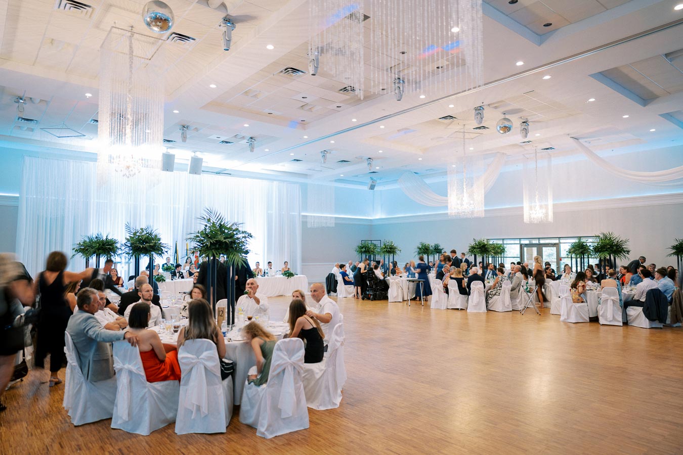 Elegant wedding reception hall with elegantly dressed guests seated at round tables, adorned with tall floral centerpieces, white chair covers, and soft lighting from chandeliers, creating a sophisticated and celebratory atmosphere.