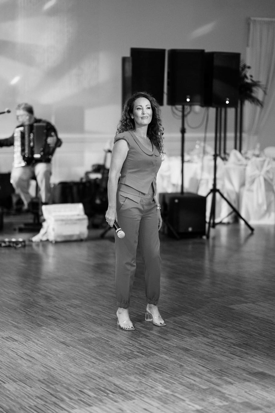 A woman in a stylish outfit holding a microphone stands confidently on a polished wooden floor, ready to perform at an indoor event. In the background, a musician plays an accordion under soft lighting, with speakers and elegantly arranged tables visible.