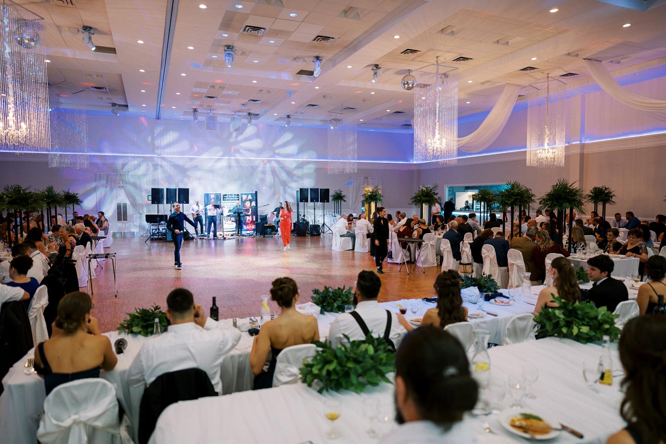 A vibrant wedding reception in a spacious hall, featuring elegantly decorated tables with guests enjoying a formal dinner. The atmosphere is lively with a dance floor and live band performing under soft lighting, surrounded by decorative plants and draped ceiling fabric.