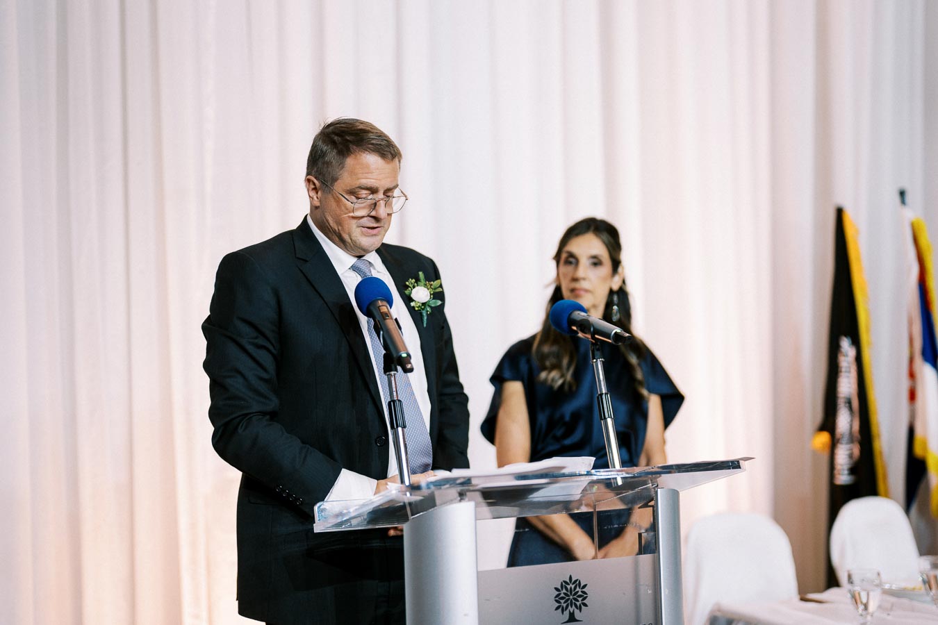 A man and a woman stand at a podium speaking into microphones during a formal event, with white curtains and flags in the background. The man wears a suit with a floral boutonniere, and the woman is in a dark dress, as they appear to address an audience.