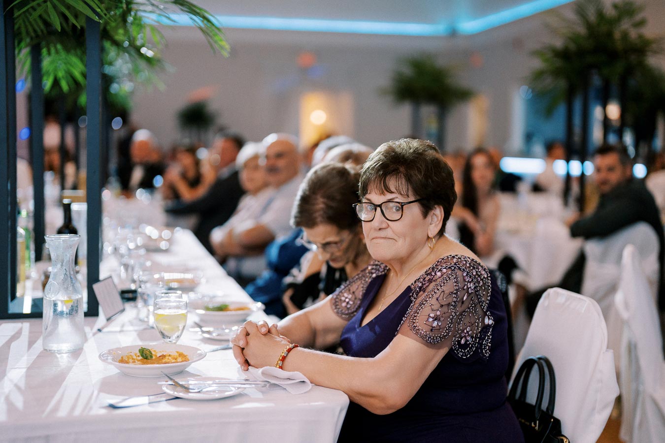 Senior woman in a purple dress sitting at an elegantly set table during a formal event, surrounded by guests in a well-lit banquet hall with green plants in the background.