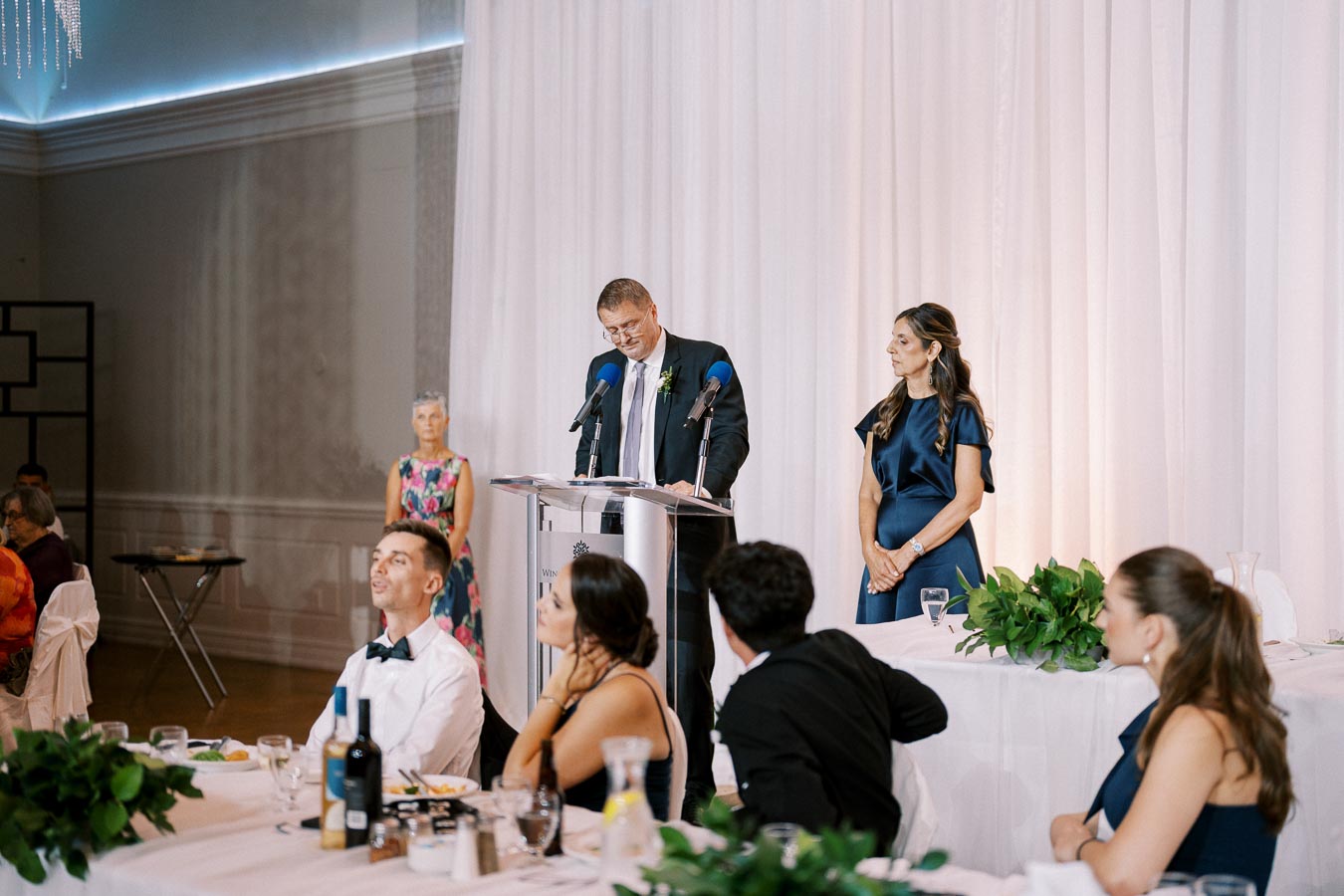 A man in a suit giving a speech at a formal event, with guests seated and listening attentively. The setting includes elegant decor with white curtains and floral arrangements.
