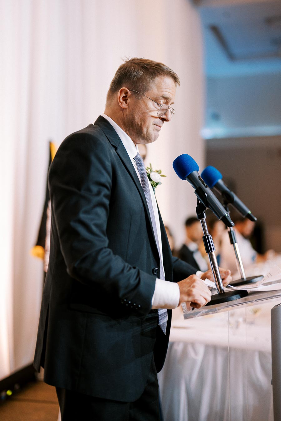 Man delivering a speech at a podium with two microphones, wearing a suit and tie, and a flower boutonniere, set against a softly lit background.