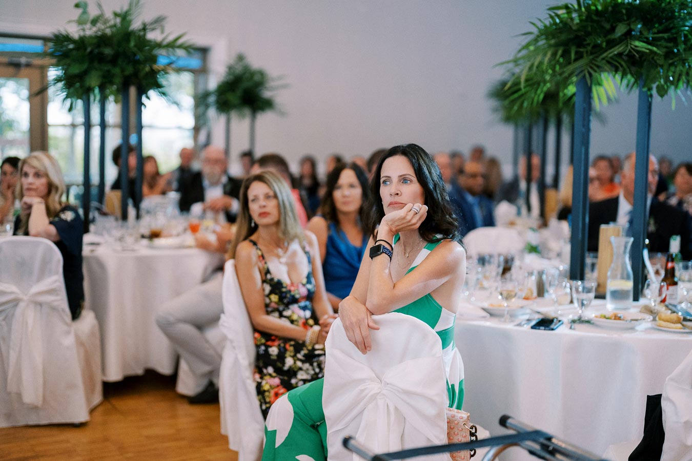 Guests attentively listening at a formal event, with elegantly set tables and lush greenery decor in the background, creating a sophisticated atmosphere.