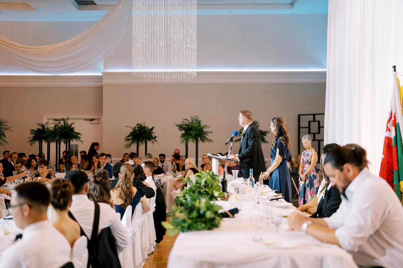 Wedding reception with guests seated at tables, greenery decor, and a speaker in a suit addressing the audience from a podium in a decorated banquet hall.