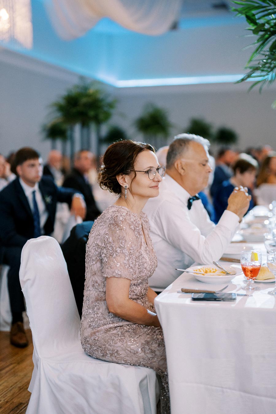 Guests sitting at elegantly set dining tables during a formal event, with a woman in a lace dress attentively watching the proceedings.